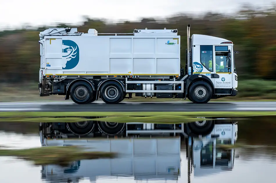 A white rubbish lorry with green and blue logos drives quickly along a road, with its reflection visible in a pool of water below. Trees and grass are blurred in the background, emphasising the lorry's motion.
