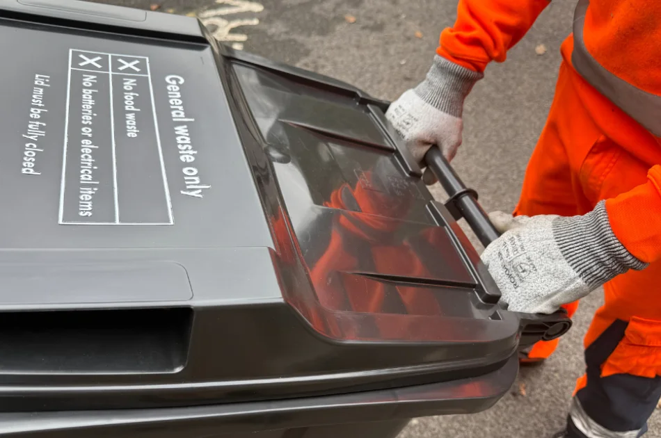 A person wearing orange work clothes and protective gloves is moving a black general waste wheelie bin with a closed lid on a paved surface.