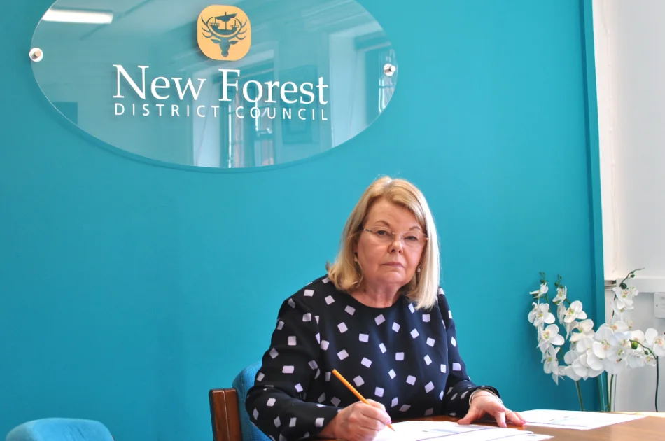 A woman with blonde hair and glasses wearing a dark polka-dot top sits at a desk writing, with a New Forest District Council sign on a teal wall behind her and white flowers on the table.