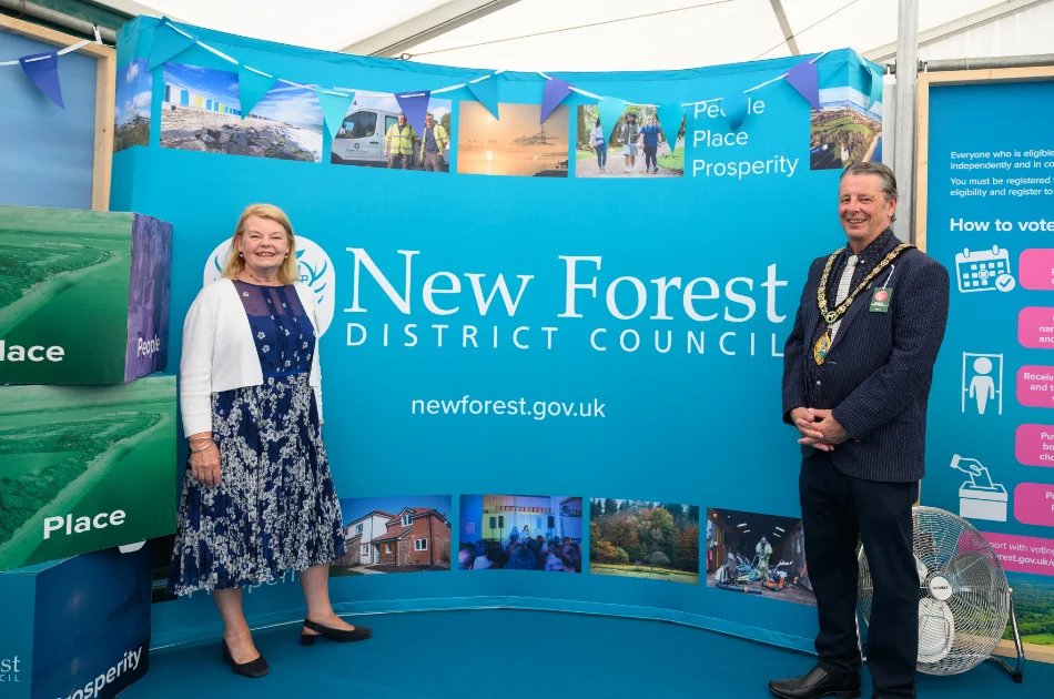 Two people stand in front of a New Forest District Council display with photos and the words “People, Place, Prosperity.” They are smiling, and there are large cubes labelled “Place” beside them.