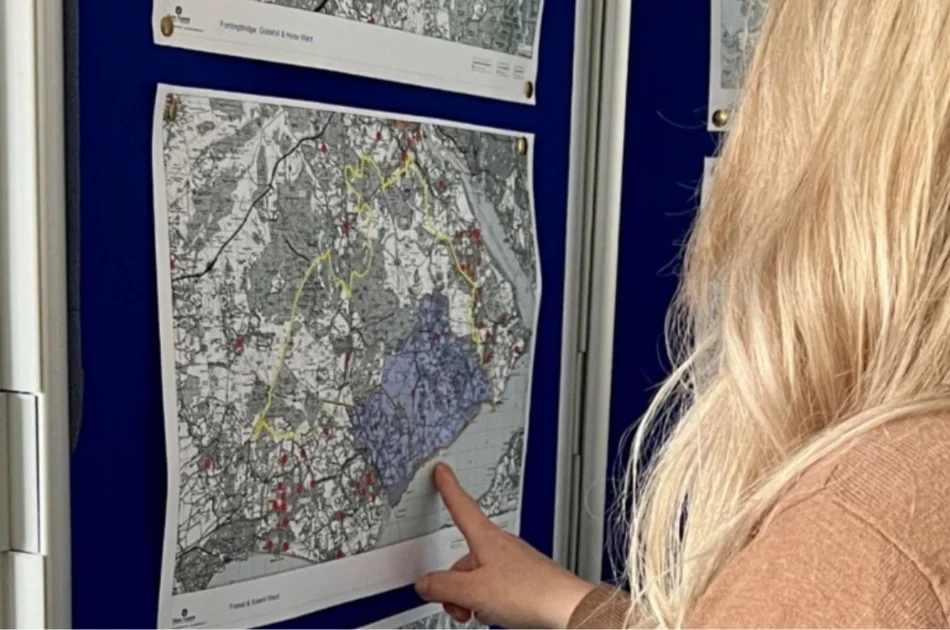 A person with long blonde hair points to a specific area on a detailed, black-and-white map pinned to a blue display board. Other maps are also visible on the board.