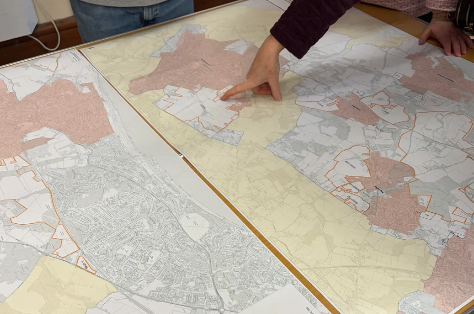 Two people are examining detailed maps on a table; one person points to a specific area on the central map, which shows bordered regions in various shades of colour.